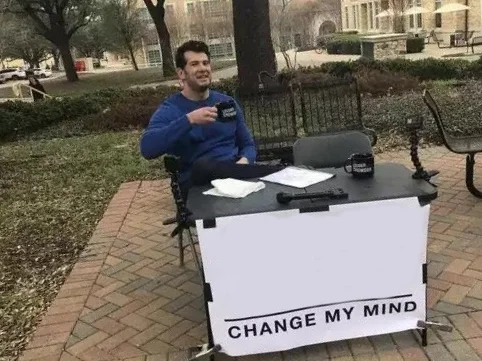 Change My Mind meme template: a man sitting at a folding table behind a handwritten sign inviting debate